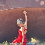 Leyya Mona Tawil is sitting on the ground in a red dress, holding up one arm.