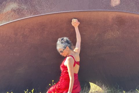 Leyya Mona Tawil is sitting on the ground in a red dress, holding up one arm.