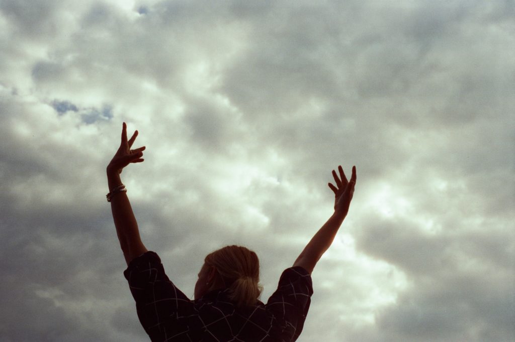 person standing against a cloudy sky, hands in the air. photo credit Chanelle Allesandre