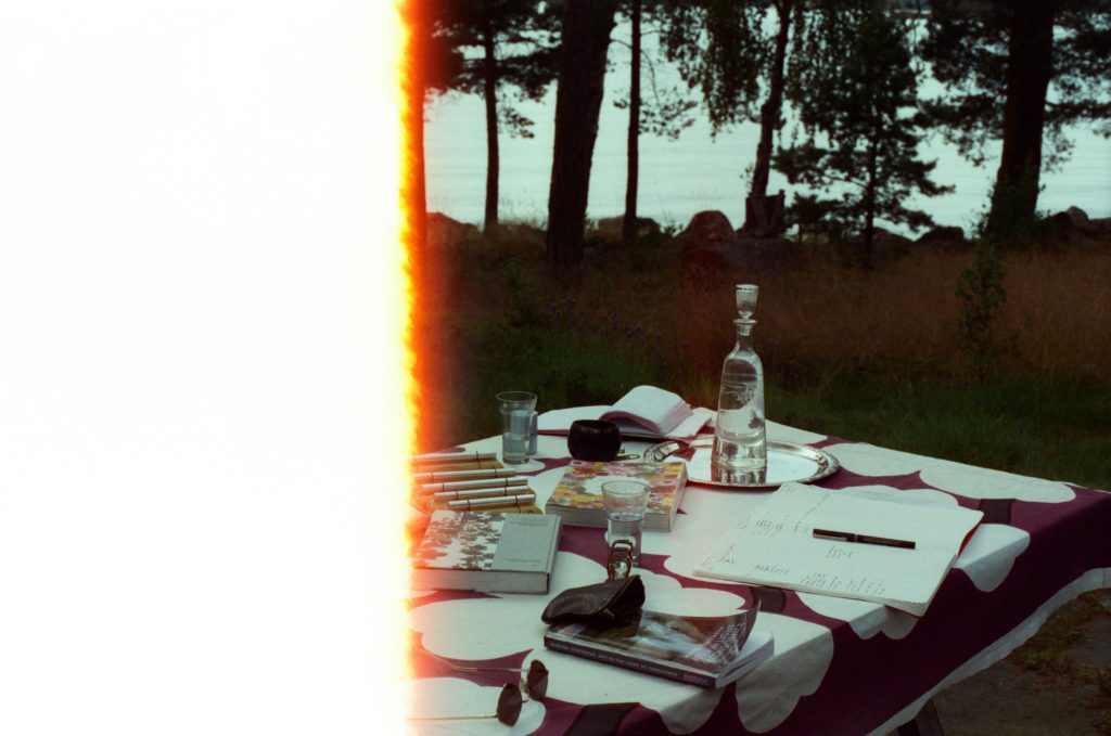 some books, notebooks, glasses and bottles on a table outside. in the background some pines, rocks and the sea. photo credit Chanelle Allesandre