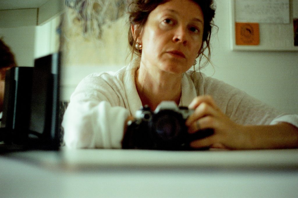 Chanelle sitting at a table in front of a mirror, holding a camera. phptp credit Chanelle Allesandre.