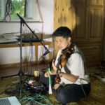 Andrea Lim is sitting on the floor playing a violin, speaking into a microphone surrounded by cables and effect pedals.