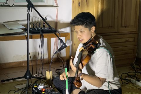 Andrea Lim is sitting on the floor playing a violin, speaking into a microphone surrounded by cables and effect pedals.