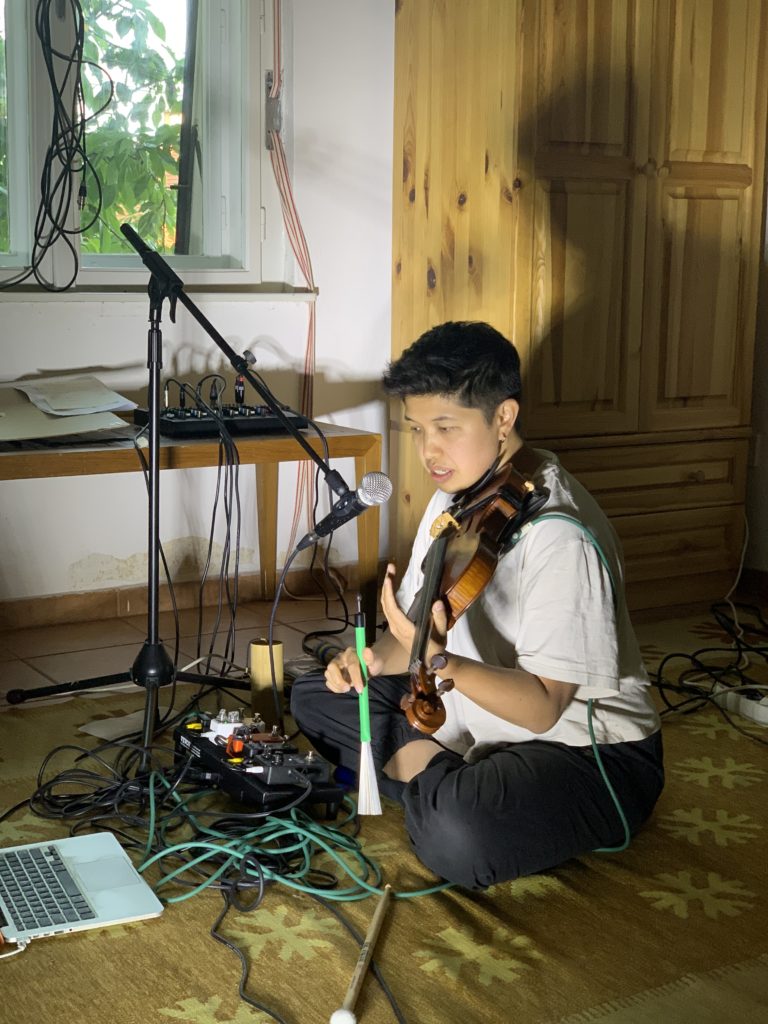 Andrea Lim is sitting on the floor playing a violin, speaking into a microphone surrounded by cables and effect pedals.