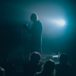 photo of a human holding a microphone in a dark room, surrounded with spectators and a dim blue light