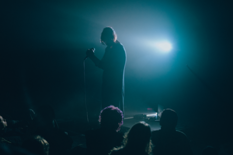 photo of a human holding a microphone in a dark room, surrounded with spectators and a dim blue light