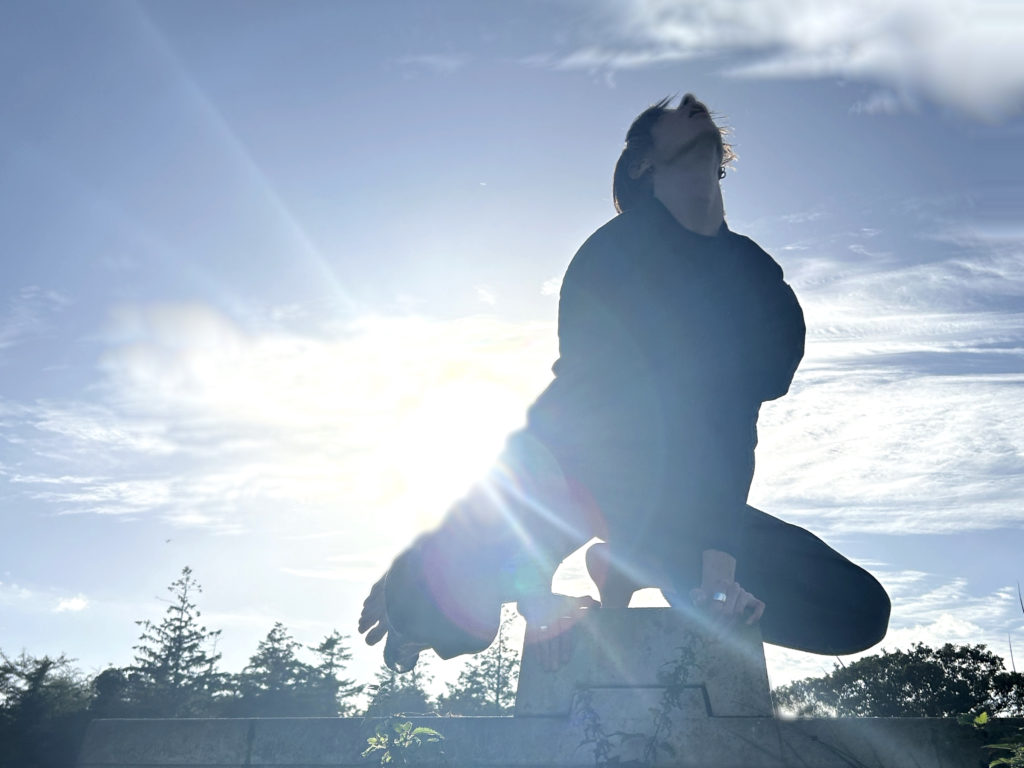 person looking up into the sky in a squat position, with the sun and some forest behind them