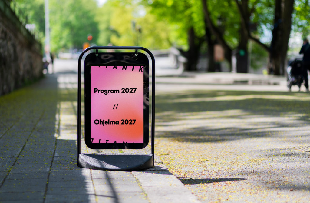 Kadulla on ständi, jossa on vaaleanpunainen juliste. Julisteessa lukee: Program 2027 / / Ohjelma 2027.