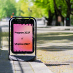 Kadulla on ständi, jossa on vaaleanpunainen juliste. Julisteessa lukee: Program 2027 / / Ohjelma 2027.