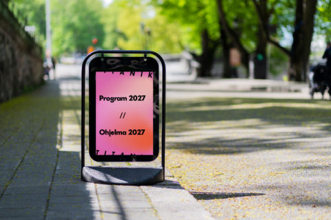 Kadulla on ständi, jossa on vaaleanpunainen juliste. Julisteessa lukee: Program 2027 / / Ohjelma 2027.
